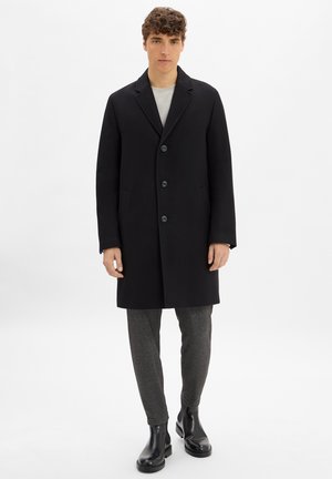 Schwarzer Wollmantel mit Reverskragen, vorderem Knopfverschluss und Seitentaschen. Getragen über einem grauen Hemd und grauen gemusterten Hosen. Schwarze Stiefel.