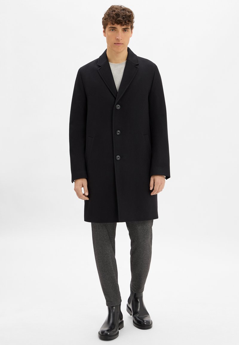 Cappotto di lana nera con colletto a scollo a V, chiusura anteriore con bottoni e tasche laterali. Indossato sopra una camicia grigia e pantaloni grigi a motivi. Stivali neri.