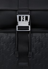 Must tauer messenger kott, millel on tekstureeritud geomeetriline muster, hõbedased riistade kinnitused koos logoga ja kudumise detailidega rihm.