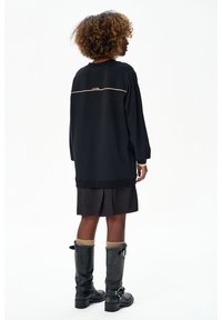 Osoba o kręconych włosach ubrana w czarną, oversize'ową bluzę, czarną plisowaną spódnicę, beżowe skarpetki i czarne kozaki za kolano, stoi tyłem.