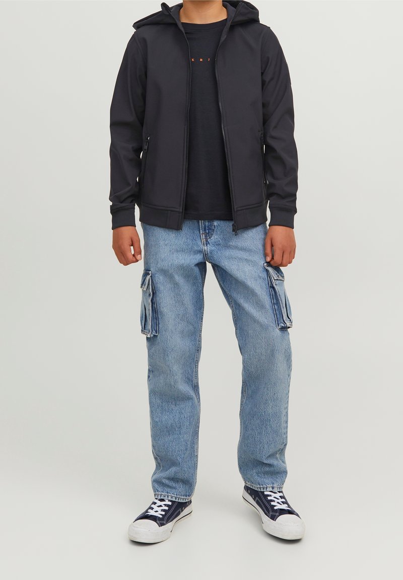 Personne portant une veste à capuche noire zippée, un t-shirt noir, un jean cargo bleu clair et des baskets noires et blanches, debout devant un fond neutre.