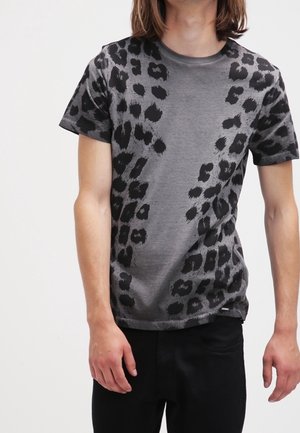 Personne portant un t-shirt gris à manches courtes avec des taches léopard abstraites noires et un pantalon noir, debout devant un fond uni.