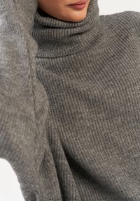Szary sweter z golfem wykonany z dzianiny z prążkowaną fakturą, z wysokim kołnierzem i luźnym krojem. Materiał wydaje się miękki i ciepły.