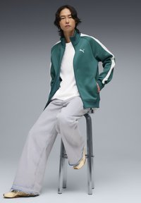 Πράσινο αθλητικό μπουφάν με λευκά στοιχεία, συνδυασμένο με ανοιχτό γκρι φαρδιά τζιν και κίτρινα παντόφλες, καθισμένος σε σκαμπό σε γκρι φόντο.