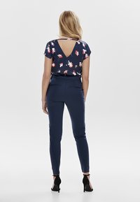 Marineblauwe bloes met bloemenpatroon en een V-uitsnijding aan de achterkant, gecombineerd met slim fit marineblauwe broek. Soepele textuur, aangevuld met zwarte hoge hakken.