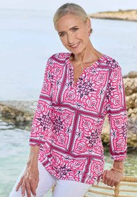 Langsleeve, roze gestippeld top met bloemdesigns en contrasterende zwarte accenten, gecombineerd met witte broek, tegen een kustachtergrond.