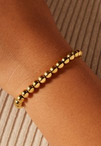 Gouden armband met kralen in een glad en afgerond ontwerp. Voorzien van gelijkmatig verdeelde metalen kralen die glanzen tegen een huidkleurige pols.