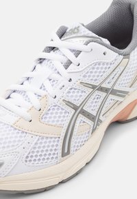 Sapatilha atlética branca com parte superior em malha, detalhes cinzentos e sola de borracha. Apresenta um design em camadas com detalhes em bege e cinza.