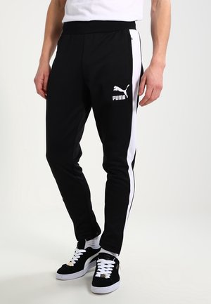 Mand iført sorte Puma træningsbukser med hvide sidelister og sorte Puma-sneakers med hvide snørebånd, stående mod en ensfarvet baggrund.