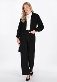 Černý dlouhý cardigan vyrobený z měkkého materiálu, s volnějším střihem, otevřeným předním dílem a dvěma předními kapsami, v kombinaci s bílou košilí a černými kalhotami.