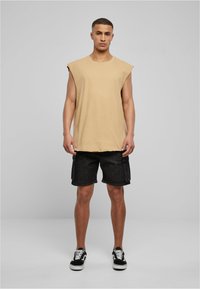 Beige mouwloos T-shirt van zachte stof, gecombineerd met zwarte denim cargoshorts met zijzakken en een relaxed fit, gedragen met zwarte sneakers.