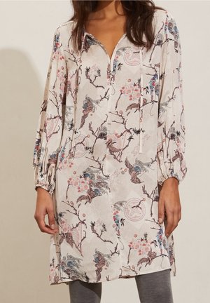 Donna che indossa una tunica a maniche lunghe con un motivo floreale e di uccelli in toni morbidi di rosa, blu e marrone sopra leggings grigi.