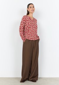 Bunte rot-creme gemusterte Bluse mit V-Ausschnitt und langen Ärmeln, kombiniert mit lockeren braunen Hosen mit weitem Bein. Glatte Stoffstruktur.