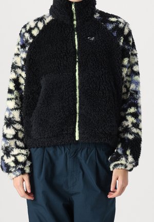 Schwarze Fleecejacke mit gemusterten schwarzen, weißen und blauen Ärmeln, Frontreißverschluss und elastischer Taille. Dunkelblaue Hose sichtbar.