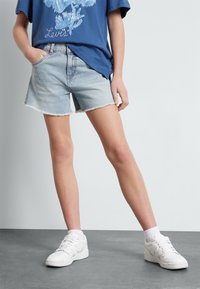 Jasnoniebieskie szorty z denimu z postrzępionym wykończeniem, sięgające do połowy uda, prezentowane z niebieskim T-shirtem z grafiką i białymi trampkami. Prosty design, casualowy styl.