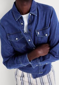 Denim shirt in dark blue with two chest pockets, button-up front, and contrasting stitching. Layered over a white ribbed shirt.