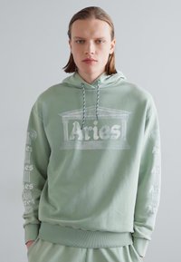 Haut à capuche vert clair avec un graphique sur le devant représentant un design de colonne classique et le texte "Aries". Manches longues avec le texte répétitif "Aries".