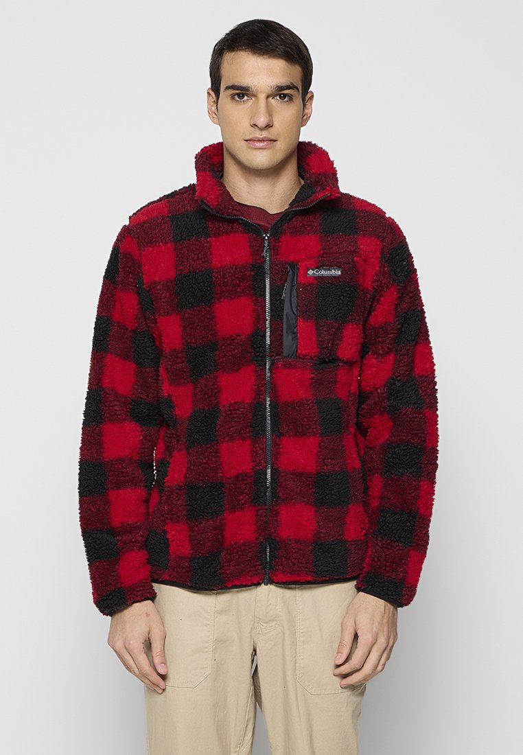 Młody mężczyzna w czerwoną i czarną kraciastą polarową kurtkę z zamkiem z przodu i kieszenią na piersi, stojący na tle jasnego, jednolitego tła.