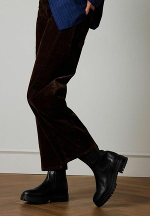 Persoon draagt donkerbruine corduroy broek en zwarte leren enkellaarsjes, staat op een houten vloer tegen een effen muur.