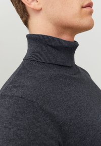 Gros plan sur une personne portant un pull col roulé côtelé gris foncé, montrant le cou et une partie de l'épaule sur un fond neutre.