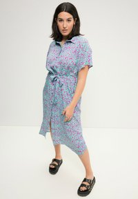 Abito camicia a maniche corte di colore azzurro chiaro con motivo astratto viola, legato in vita, con un orlo spaccato e abbinato a sandali neri.