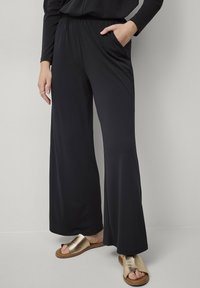 Zwarte wijde broek gemaakt van zacht, rekbaar materiaal met een elastische tailleband en zijzakken, gecombineerd met metallic slip-on sandalen.
