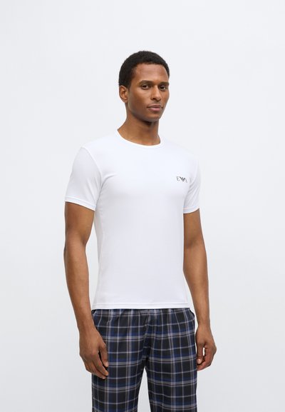 Bílé tričko s krátkým rukávem a kulatým výstřihem s malým vyšívaným logem v horní levé části. Nošeno s modro-černými károvanými pyžamovými kalhotami.