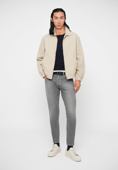 Μπεζ ελαφρύ μπουφάν με ελαστικούς καρπούς, ναυτικό πουκάμισο από κάτω, γκρι slim-fit τζιν και κρεμ αθλητικά παπούτσια. Το μοντέλο στέκεται μπροστά σε λευκό φόντο.