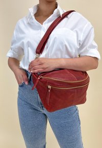 Borsa a spalla in pelle rossa con due scomparti con cerniera, una tracolla regolabile e una texture liscia, esposta su uno sfondo neutro.
