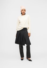 Γυναίκα που φοράει ελαφρύ ριγέ πουλόβερ, ροζ σκουφάκι, μαύρη πλισέ φούστα από τζιν πάνω από μαύρο παντελόνι και μαύρες μυτερές γόβες, στέκεται μπροστά σε λευκό φόντο.