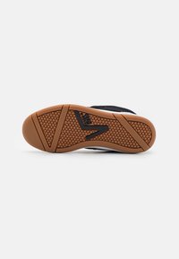 Semelle de chaussure avec une semelle extérieure texturée marron, logo noir au centre et empeigne en mesh noire visible sur les bords sur un fond blanc uni.