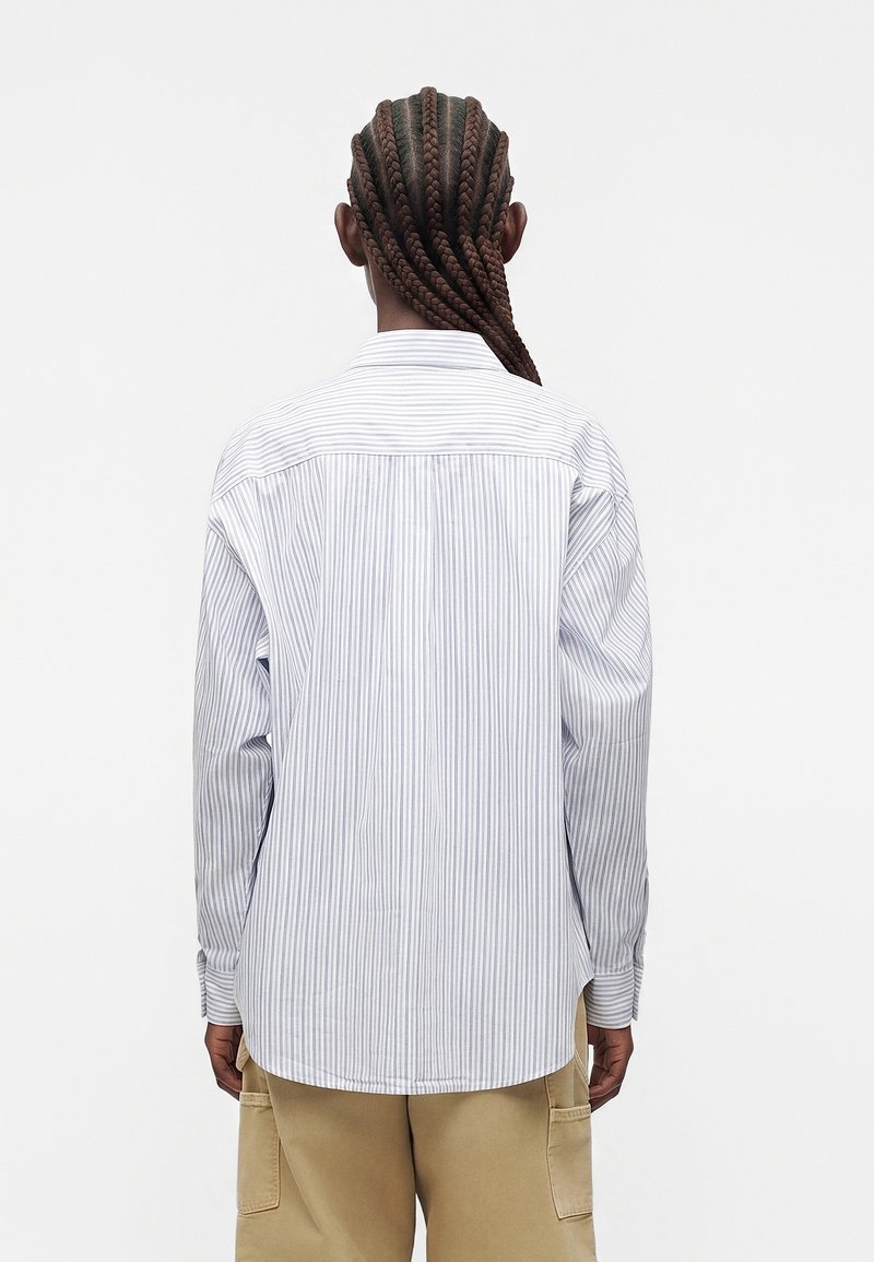 Persona con lunghi capelli intrecciati indossa una camicia a maniche lunghe a righe azzurro chiaro e pantaloni beige, in piedi di spalle contro uno sfondo semplice.