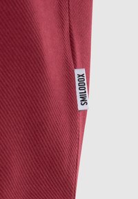 Tessuto rosso testurizzato con un motivo diagonale; include un'etichetta bianca con scritto "SMILODON." Focalizzato su un materiale liscio e durevole.