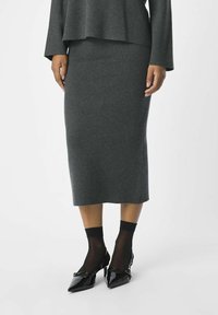 Grå stickad midikjol med en åtsittande form, i kombination med svarta lackade pumps med spetsig tå och genomskinliga svarta strumpor som slutar vid anklarna.