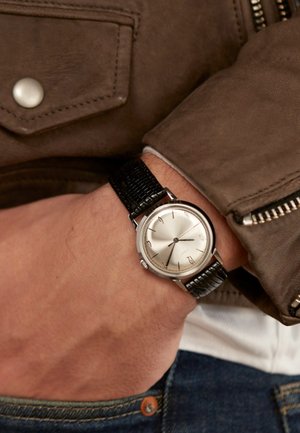 Montre en argent avec un cadran rond, bracelet en cuir de crocodile noir, marqueurs d'heure minimalistes et un cadran en argent texturé. Portée au poignet.