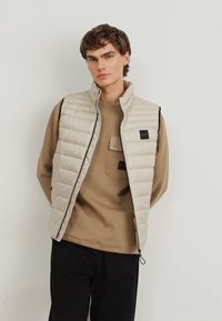 Hellbeige gesteppte Weste mit hohem Kragen, schwarzem Besatz und Logo-Patch, getragen über einem hellbraunen Langarmshirt. Schwarze Hose vervollständigt das Outfit.