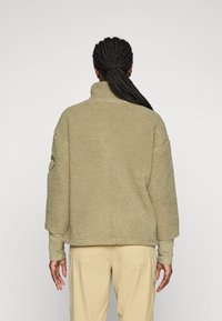 Persona con capelli intrecciati che indossa una giacca in pile beige chiaro e pantaloni beige, mostrata di spalle su uno sfondo semplice.