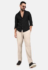 Camicia nera con bottoni e finish testurizzato, pantaloni beige larghi e sneakers nere. Gli accessori includono occhiali da sole e una catena.