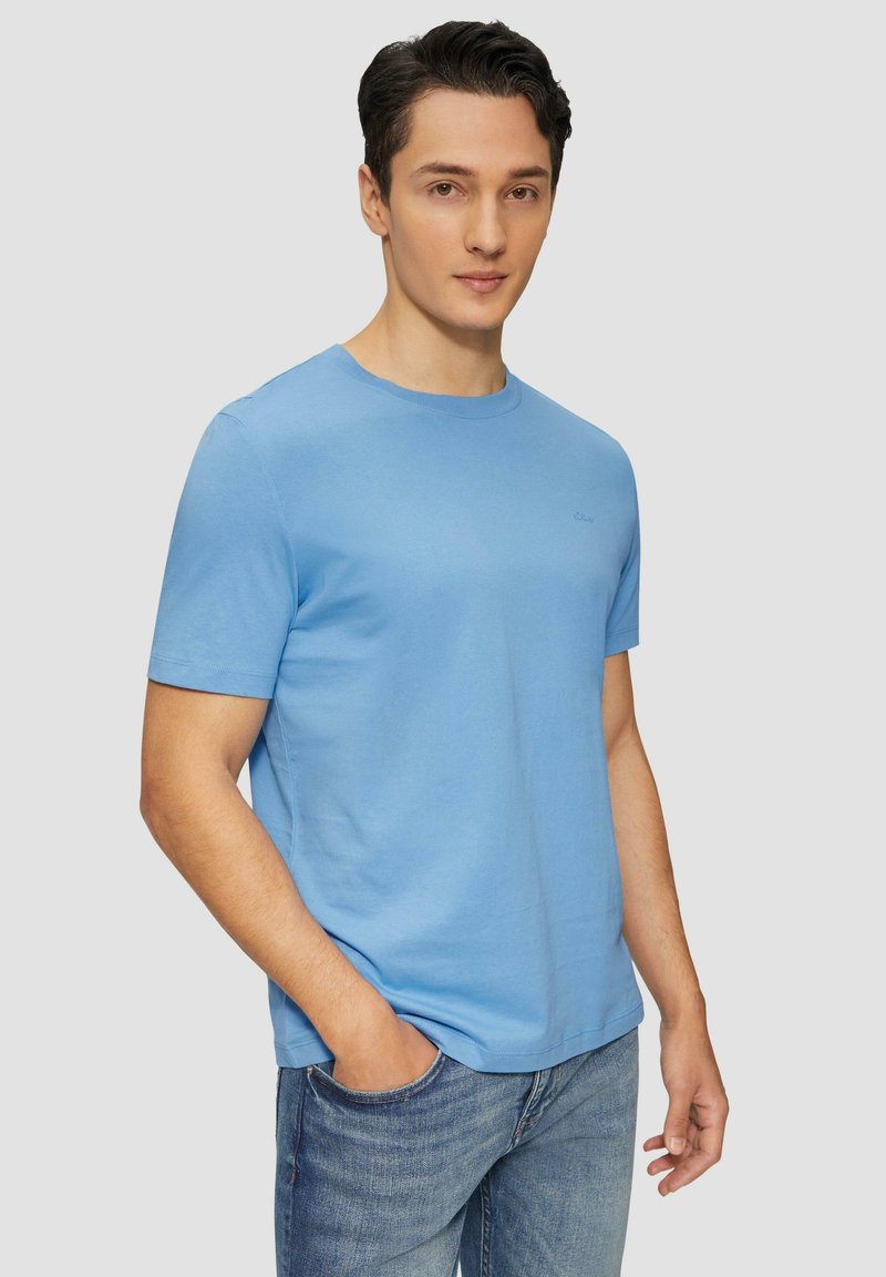 Jasnoniebieska bawełniana koszulka z okrągłym dekoltem, krótkimi rękawami i subtelnym detalem logo na klatce piersiowej. Noszona z niebieskimi dżinsami.