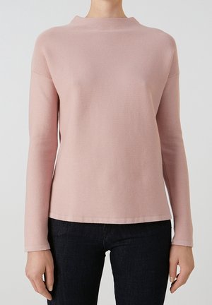 Kvinde iført en lyserød langærmet top med bådudskæring og mørkeblå tætsiddende jeans foran en ensfarvet grå baggrund.