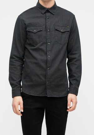 Homme portant une chemise en denim noire à boutons avec des poches à rabats sur la poitrine, associée à un pantalon noir, debout devant un fond clair uni.