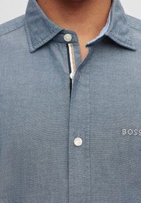 Camicia di jeans azzurro chiaro, con colletto a punta, bottoni bianchi e un delicato motivo testurizzato con dettagli a righe a contrasto all'interno della patta.