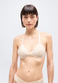 Γυναίκα με κοντά σκούρα μαλλιά που φοράει μπεζ δαντελένιο bralette και εσώρουχα που ταιριάζουν, στραμμένη προς τα εμπρός σε ένα απλό ανοιχτό φόντο.