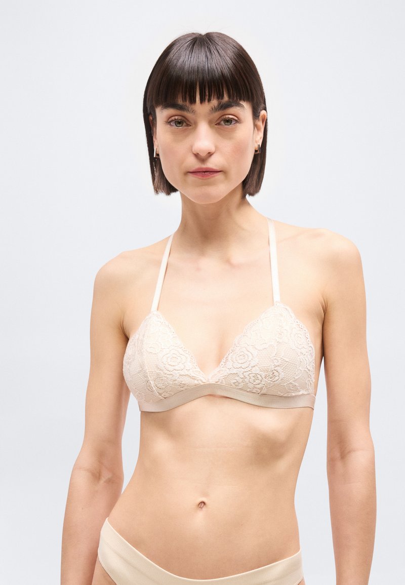 Γυναίκα με κοντά σκούρα μαλλιά που φοράει μπεζ δαντελένιο bralette και εσώρουχα που ταιριάζουν, στραμμένη προς τα εμπρός σε ένα απλό ανοιχτό φόντο.