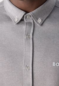 Grijze textuur knoopshirt met een kleine kraag, doorzichtige knopen en een merklogo aan de rechter onderzijde.