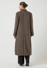 Abrigo largo marrón con acabado texturizado, que cuenta con una abertura trasera, un cuello y puños con botones. Combinado con vaqueros oscuros y zapatos negros.