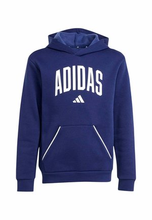 Ragazzo che indossa una felpa blu navy Adidas con logo bianco e pantaloncini grigi Adidas, in piedi davanti a uno sfondo chiaro uniforme.