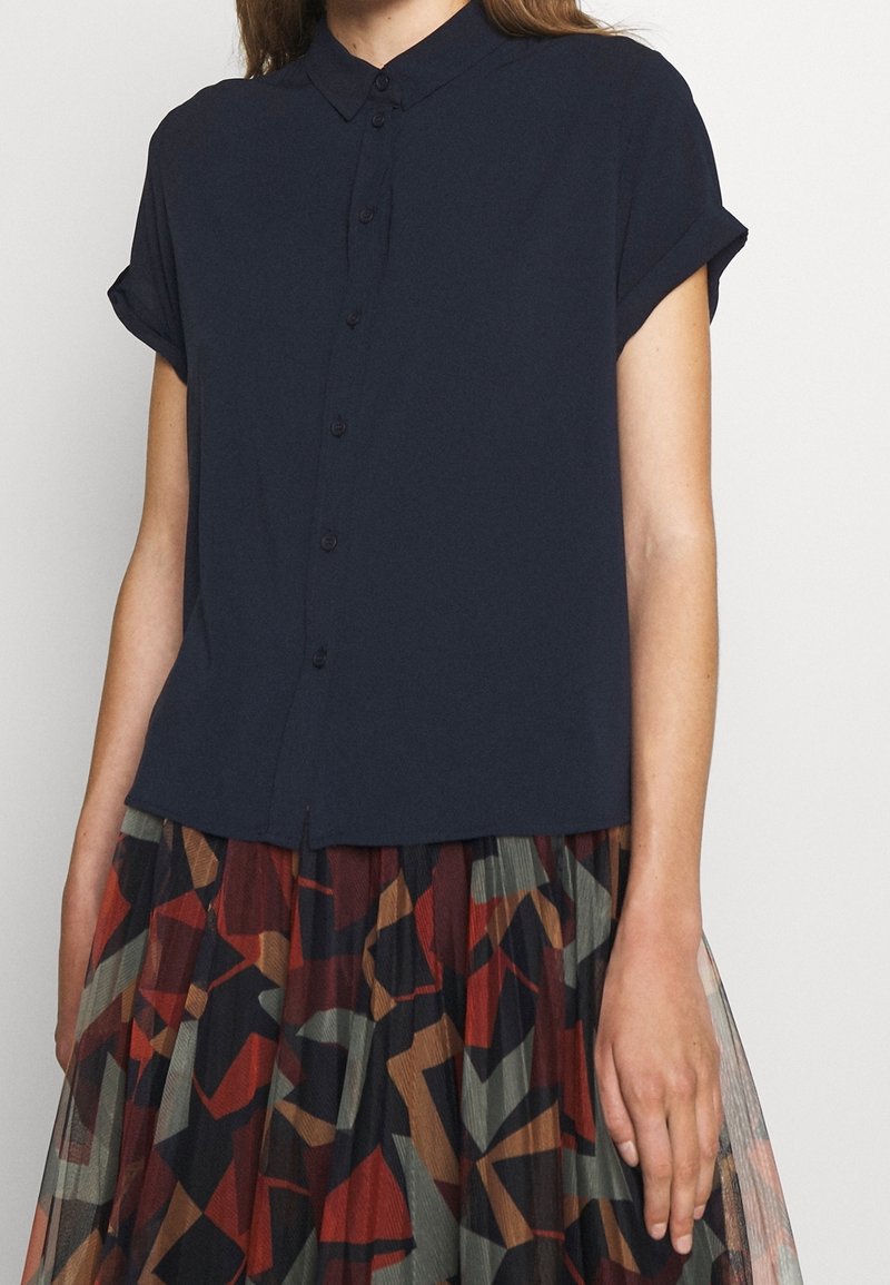 Femme portant une chemise bleu marine à manches courtes avec boutons et une jupe plissée aux motifs géométriques abstraits rouges, marron, noirs et verts.