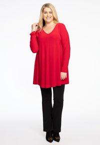 Rode langemouwen tuniek met een V-hals, losse pasvorm die onder de heupen valt, gecombineerd met zwarte fitted broek en zwarte hoge hakken.