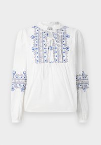Chemisier blanc avec broderies bleues et noires le long du devant et des manches. Présente un décolleté à nouer et un corps froncé. Manches longues élastiques.